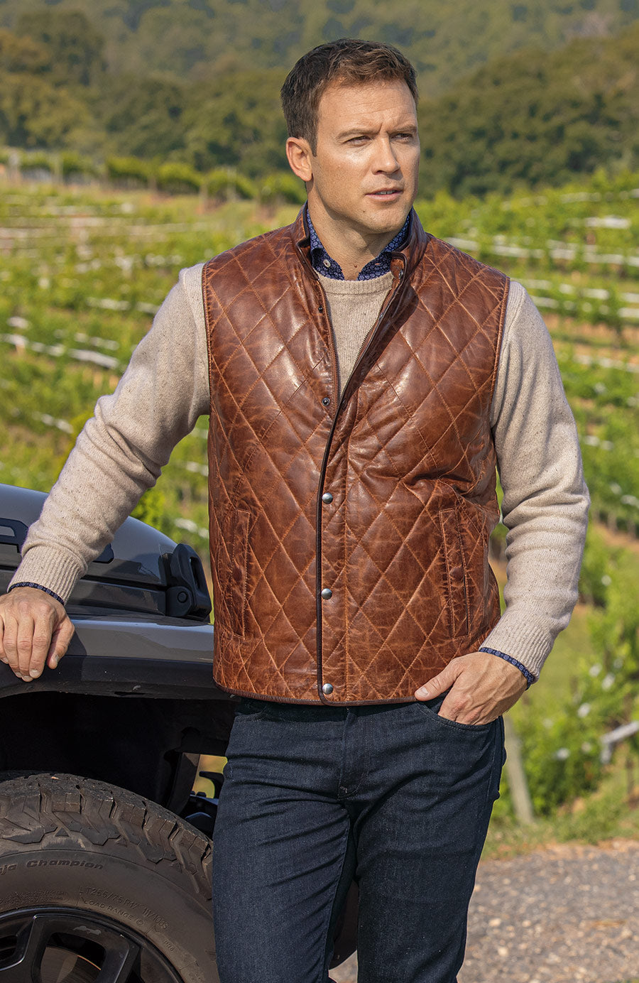 male model wearing leather vest standing in front of vineyard